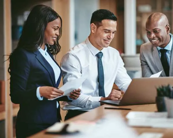 Three colleagues standing at a desk, one holding a printed report, all three looking at a laptop screen together.